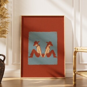 May include: A rectangular wall art print featuring a terracotta red background with a central blue square. The illustration depicts two figures in cowboy hats sitting back-to-back, wearing long-sleeved shirts and trousers, holding a small brown jug.