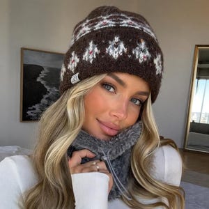 May include: A dark brown knit beanie featuring a white and red geometric fair isle pattern and a small white tag with the text LET. The beanie is paired with a dark grey faux fur neck warmer that includes a clear plastic drawstring toggle.