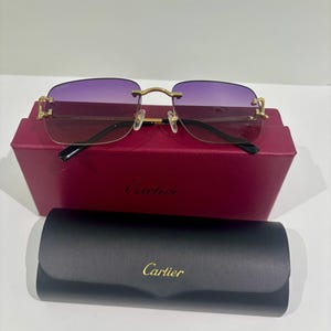 Puede incluir: Gafas de sol sin montura con lentes degradadas en color morado y herrajes de metal dorado. Las gafas descansan sobre una caja Cartier burdeos, situada detrás de un estuche rígido negro Cartier con el nombre de la marca en letras doradas.