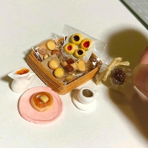 以下が含まれることがあります： 小さなプラスチック包装されたクッキーやペストリーが入った茶色の編みかご、白い陶器のピッチャー、バターをのせたパンケーキがのったピンクの皿、ソーサー付きの白いコーヒーカップで構成されたミニチュアセット。
