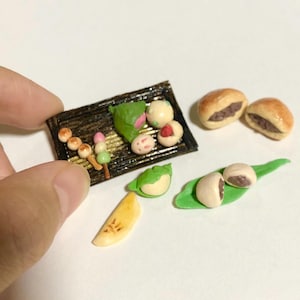 以下が含まれることがあります： 小さな団子の串や丸い菓子がのった暗い長方形のトレイ、そして金色の焼き菓子2つと緑の葉の上に置かれた白い丸い菓子2つを含む、ミニチュアフードのコレクションです。