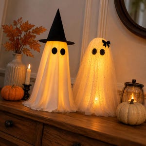 May include: Two illuminated ghost decorations. One ghost wears a black witch hat and has a smooth white fabric body. The second ghost is draped in white glittery tulle fabric with a small black bow. Both have round black eyes and glow from within.