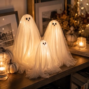 Puede incluir: Tres decoraciones con forma de fantasma hechas de tela blanca transparente drapeada sobre estructuras iluminadas. Cada fantasma presenta dos ojos negros circulares y una boca negra circular más pequeña. Las figuras emiten un brillo cálido.