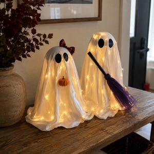 Op de afbeelding: Twee witte spookdecoraties gedrapeerd in stof en verlicht door interne lichtsnoeren. Het ene spook heeft een kleine oranje pompoen en een strik met patroon, terwijl het andere een zwarte bezemsteel met paarse borstelharen vasthoudt. Zwarte ogen.