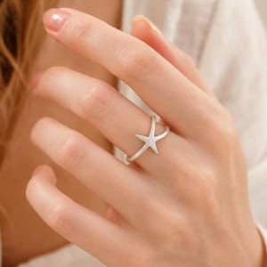 Puede incluir: Un anillo de color plateado con un delicado diseño de estrella de mar. La banda es fina y lisa, con el motivo de la estrella de mar posicionado en el centro. El metal tiene un acabado mate y cepillado, creando una apariencia sutil y discreta.