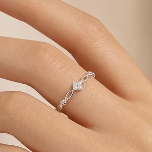 Puede incluir: Un anillo fino de plata que presenta un grupo central de pequeñas piedras claras en forma cuadrada. La banda está diseñada con delicados bucles de metal calado a ambos lados del engaste central, creando un aspecto refinado y esbelto.