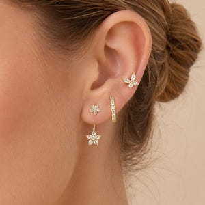 Puede incluir: Un conjunto de pendientes dorados que incluye un pequeño pendiente de botón con forma de mariposa, uno diminuto con forma de flor, un aro fino con piedras transparentes y un pendiente colgante con forma de flor y piedras transparentes.