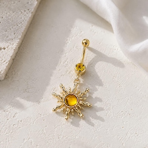 Puede incluir: Un piercing para el ombligo en tono dorado con una barra recta, un pequeño cristal amarillo y un dije en forma de sol. El dije de sol tiene una piedra amarilla central rodeada de rayos ondulados y texturizados, creando un diseño radiante.