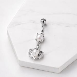 Può includere: Un piercing per l'ombelico a barra curva color argento con una piccola sfera rotonda sulla parte superiore, una perla bianca centrale fissata da quattro griffe e un ciondolo a forma di conchiglia con una perla bianca al centro.