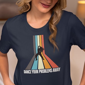 May include: A navy blue short-sleeved t-shirt featuring a graphic of a person dancing in silhouette against vertical stripes in grey, light blue, yellow, and orange. The text "DANCE YOUR PROBLEMS AWAY" is printed in white below the graphic.