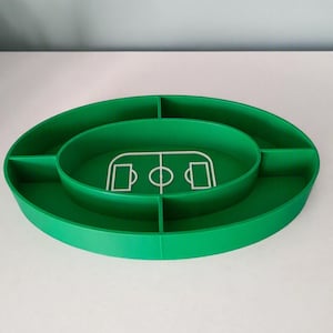 Può includere: Un vassoio da portata ovale di colore verde con quattro scomparti divisi. Il centro del vassoio presenta il contorno grafico bianco di un campo da calcio, inclusi le aree di porta, le aree di rigore e il cerchio centrale in rilievo.