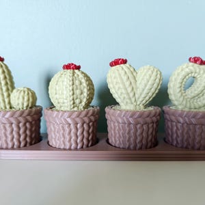 Peut inclure: Un ensemble de quatre figurines de cactus vert clair dans des pots marron à texture tricotée, disposées sur une base marron assortie. Chaque cactus est surmonté d'une petite fleur rouge, et les formes des cactus forment le mot LOVE.