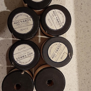May include: Six spools of white sewing thread by J. Dewhurst & Sons. Each spool features a black cap with a label stating 400 yards (366 metres) and origin details. Two spools have exposed cardboard centres, while four have printed labels on top.