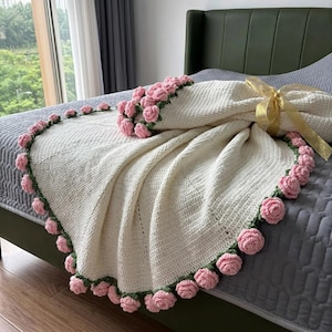 May include: A cream-colored crochet blanket featuring a textured stitch pattern and a decorative border of small, three-dimensional pink rose appliqués with green leaves. A gold ribbon is tied around the folded corner of the textile piece.