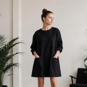 May include: A black long-sleeved mini dress featuring a round neckline and two large patch pockets on the front. The garment has a loose, relaxed fit with a straight hemline that falls above the knees, crafted from a solid dark fabric.