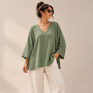 Peut inclure: Une tunique vert olive à col en V avec des manches trois-quarts et une finition en tissu texturé, associée à un pantalon large blanc cassé. L'ensemble est complété par des sandales plates marron clair et des lunettes de soleil foncées.