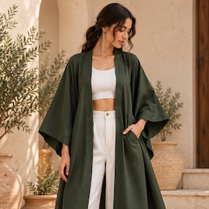 May include: A dark olive green open-front duster cardigan with wide three-quarter length sleeves and side pockets, worn over a white cropped tank top and high-waisted white wide-leg trousers with a single button closure and woven sandals.