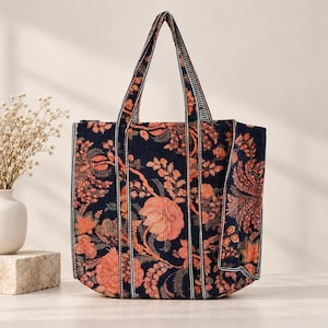 Op de afbeelding: Een gewatteerde tote bag met een donkerblauwe achtergrond en een herhalend bloemenpatroon in perzik-, oranje- en gedempte groentinten. De tas heeft twee verticaal gestreepte hengsels en een gestructureerde vorm met verticale stiksels.