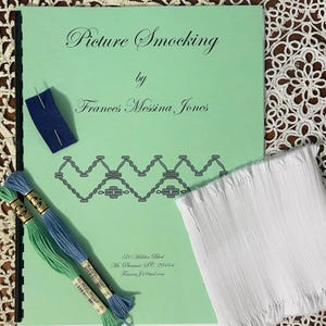 May include: A green book titled "Picture Smocking" by Frances Messina Jones. The book is open to a page with a pattern for smocking. There are two spools of embroidery floss, one blue and one green, and a piece of white fabric with a smocked edge.