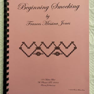 May include: A pink book with a black spiral binding titled "Beginning Smocking" by Frances Messina Jones. The book cover features a brown smocking design.