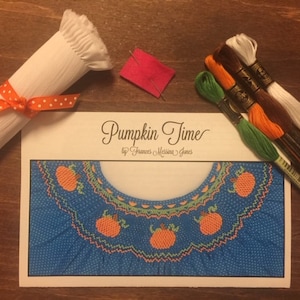 May include: A cross stitch embroidery kit featuring a blue fabric with white polka dots and a design of orange pumpkins. The kit includes white, orange, brown, and green embroidery floss, a piece of white fabric, and a pattern card that reads "Pumpkin Time by Frances Marion Jones".