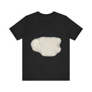 Op de afbeelding: Een zwart T-shirt met korte mouwen en een centrale afbeelding van een heremietkreeft in een lichte, onregelmatige vorm. De tekst "Changing Shells Changing Minds" is in een lichtgekleurd, schreefloos lettertype naast de illustratie gedrukt.