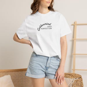 Può includere: Una t-shirt bianca a maniche corte con il testo nero "goldenmind OVER PERFECTION" stampato sul petto, abbinata a pantaloncini di jeans azzurri con orlo risvoltato e un classico design a cinque tasche con bottoni in metallo.