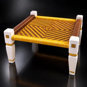 May include: A square footstool featuring four white wooden legs with gold accents. The seat is constructed from tightly woven cord in a geometric diamond pattern, alternating between vibrant yellow and deep brown colors across the surface.