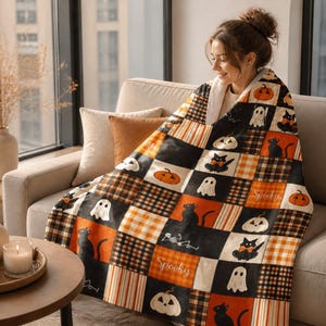 Op de afbeelding: Een patchwork-stijl plaid met een herhalend patroon van zwarte katten, witte spoken, oranje pompoenen en geruite vierkanten in oranje, zwart en wit. De deken bevat het woord Spooky gedrukt in een wit cursief lettertype.