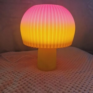 Puede incluir: Una lámpara de mesa con forma de seta que presenta una pantalla acanalada en forma de cúpula, con un degradado de rosa vibrante en la parte superior a amarillo cálido en la base. El soporte cilíndrico también es acanalado y de color amarillo.