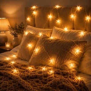 May include: A string of warm white LED star-shaped lights draped over a bed with a textured tan throw blanket, two white pillows, and a tufted headboard. The glowing star lights create a soft illumination across the bedding and the headboard.