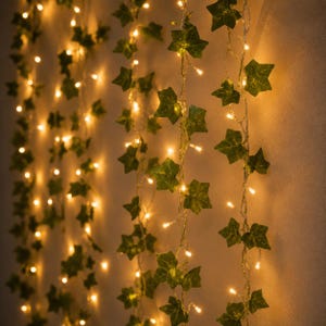 May include: Multiple vertical strands of artificial green ivy leaves intertwined with thin copper wire fairy lights. The small, warm-toned LED bulbs are spaced evenly along the vines, creating a glowing effect against the wall surface.