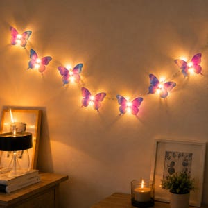 May include: A string of LED lights featuring six translucent butterfly-shaped ornaments in shades of blue and purple. Each butterfly is illuminated from within by a warm white light, creating a glowing effect against the wall surface.