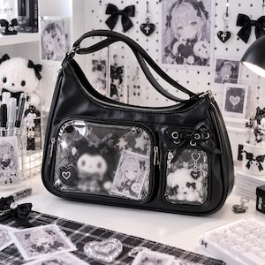 Könnte beinhalten: Eine schwarze Umhängetasche mit glatter Oberfläche, Reißverschluss oben und zwei transparenten Vordertaschen. Die größere Tasche enthält eine Plüschfigur und eine Karte, während die kleinere mit einer schwarzen Schleife und einem Plüschtier verziert ist.