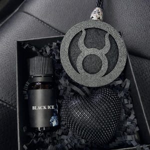 Op de afbeelding: Een zwarte geschenkdoos met een klein glazen flesje met een zwarte dop met het label BLACK ICE, een ronde zwarte hanger met het sterrenbeeld Stier en een getextureerd zwart hartvormig object in zwart versnipperd papier als vulling.