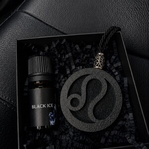 Pode incluir: Uma caixa de presente preta contendo um pequeno frasco de vidro com tampa preta rotulado como BLACK ICE e um pingente circular preto texturizado com o símbolo do zodíaco de Leão. O pingente tem um passador prateado e um cordão preto.