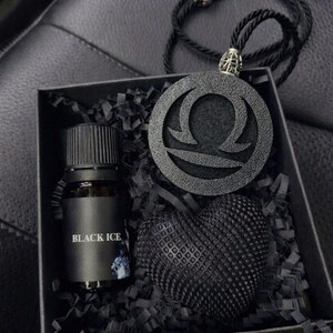 Pode incluir: Uma caixa de presente preta contendo um pequeno frasco de vidro com o rótulo Black Ice, um objeto preto texturizado em forma de coração e um pingente circular preto com o símbolo do zodíaco Libra em um cordão preto sobre papel picado.