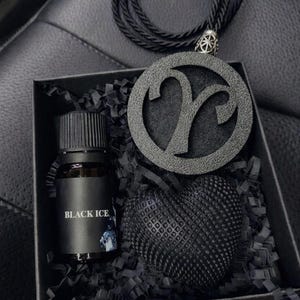 Op de afbeelding: Een zwarte geschenkdoos met een klein glazen flesje met het label BLACK ICE, een getextureerd zwart hartvormig object en een ronde zwarte hanger met een Ram-sterrenbeeld op een zwart koord, alles genest in zwart versnipperd papier.