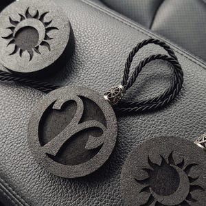 Pode incluir: Três ornamentos circulares pretos com superfícies texturizadas. Um apresenta um recorte do símbolo do zodíaco Áries, enquanto os outros dois exibem um design de sol e lua crescente. Cada peça inclui um cordão trançado preto com um passador de metal prateado.