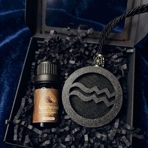 Pode incluir: Uma caixa de presente preta contendo um pequeno frasco de vidro castanho de óleo essencial de Agarwood e um pingente circular preto com o símbolo do zodíaco Aquário texturizado. O pingente está preso a um cordão preto sobre papel de enchimento.