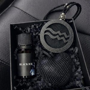Op de afbeelding: Een zwarte geschenkdoos met een klein glazen flesje met een zwart etiket met de tekst BLACK ICE, een ronde zwarte hanger met een golvend lijnontwerp aan een zwart koord, en een getextureerd zwart hartvormig object in zwart opvulmateriaal.