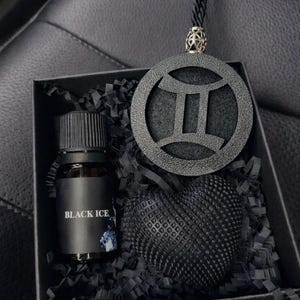 Pode incluir: Uma caixa de presente preta contendo um pequeno frasco de vidro com o rótulo BLACK ICE, um pingente circular preto com o símbolo do zodíaco de Gêmeos em um cordão preto e um objeto preto texturizado em forma de coração sobre papel.