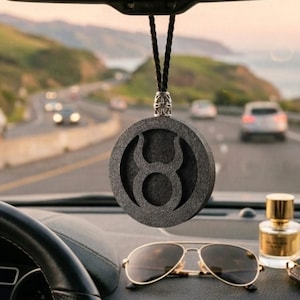 Pode incluir: Um ambientador de carro circular preto pendurado no espelho retrovisor, apresentando um recorte do símbolo do zodíaco Touro. O pingente tem uma superfície texturizada e é suspenso por um cordão trançado preto com um conector de metal prateado.