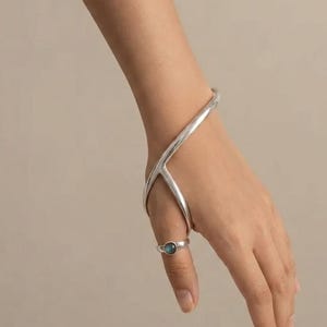 May include: A silver hand bracelet featuring a smooth, curved metal frame that extends from the wrist to the thumb, paired with a matching silver ring set with a small, round, iridescent blue-green stone centred on the thumb band.