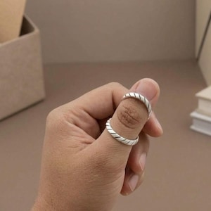 May include: A silver ring featuring a continuous diagonal grooved pattern along the band. The metal has a polished finish with dark recessed lines that create a textured, striped appearance around the circumference of the circular jewelry piece.
