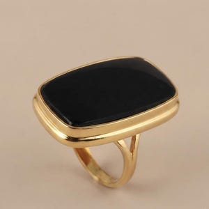 May include: A gold-toned ring featuring a large, rectangular, flat black stone set within a smooth, polished metal bezel. The band is thin and connects to the base of the setting, creating a bold, geometric look with a high-contrast color palette.