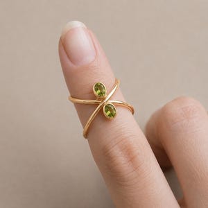 Op de afbeelding: Een gouden ring met een gekruist bandontwerp en twee ovaalvormige, lichtgroene peridoot edelstenen in individuele zettingen. De ring zit om een vinger en toont de dunne, gepolijste metalen band die de stenen op hun plek houdt.