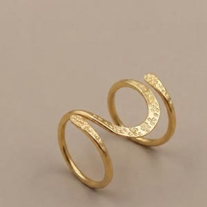 Könnte beinhalten: Ein goldfarbener Ring mit offenem Bypass-Design, bei dem sich zwei parallele Bänder zu einer zentralen S-Form krümmen. Die Oberfläche des Metalls weist eine subtile, gehämmerte Textur auf, die für ein reflektierendes und unebenes Finish sorgt.