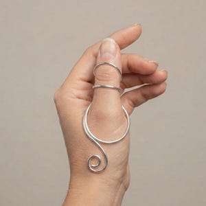 May include: A silver-colored metal finger splint featuring two circular bands that wrap around the thumb and a decorative spiral extension that curves downward along the side of the thumb, providing a structured and metallic appearance.