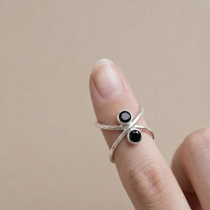 May include: A silver crossover ring featuring two round, faceted black stones set in bezel settings. The band consists of two thin, polished silver wires that intersect to create an open, bypass-style design, showcasing a sleek metallic finish.